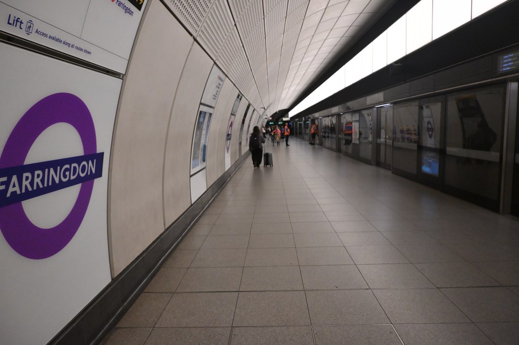 Interior of Farringdon Station, showing the platform with a purple London Underground roundel and passengers walking along the tiled walkway.