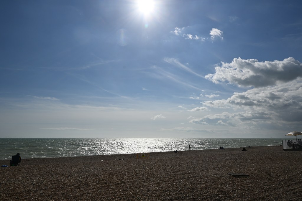 A sunny beach scene with a sparkling sea and scattered clouds under a bright blue sky. People are seen enjoying the beach, with a few sitting on the pebbled shore.