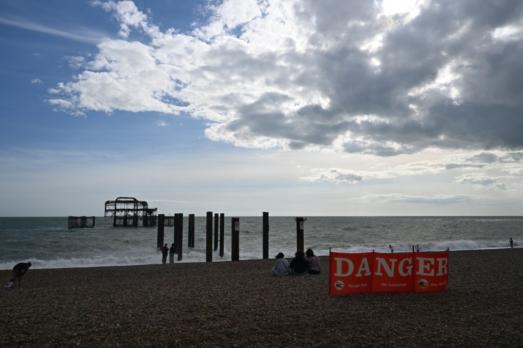 A beach scene in Brighton, showing a warning sign stating 'DANGER' in bold letters. The beach is pebbly with people sitting and walking nearby, while an old pier structure can be seen in the background under a cloudy sky.