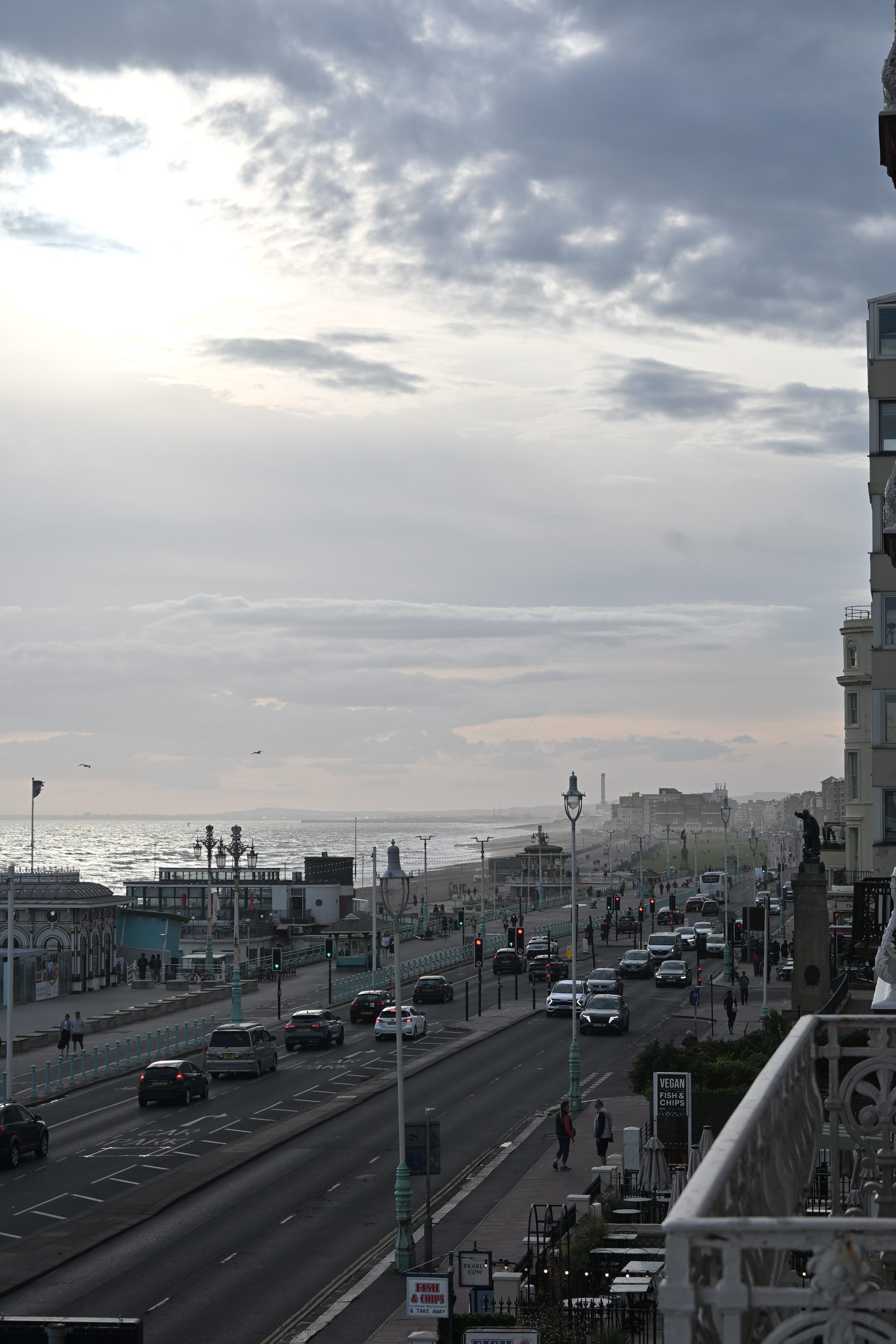 View of a coastal street in Brighton, UK, with cars, pedestrians, and the sea in the background under a cloudy sky.