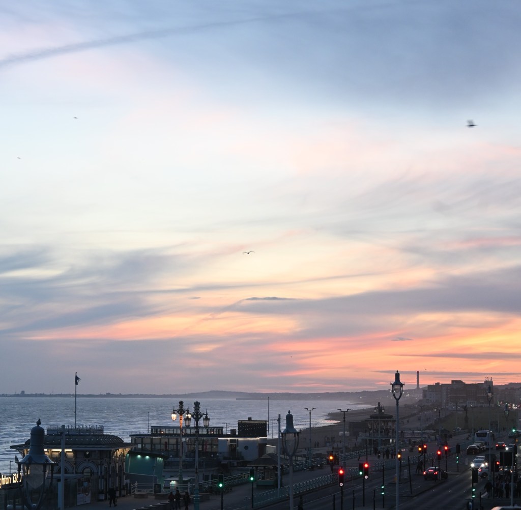A sunset view over the sea, with soft pastel colours in the sky and silhouettes of a beachfront promenade and buildings in the foreground.
