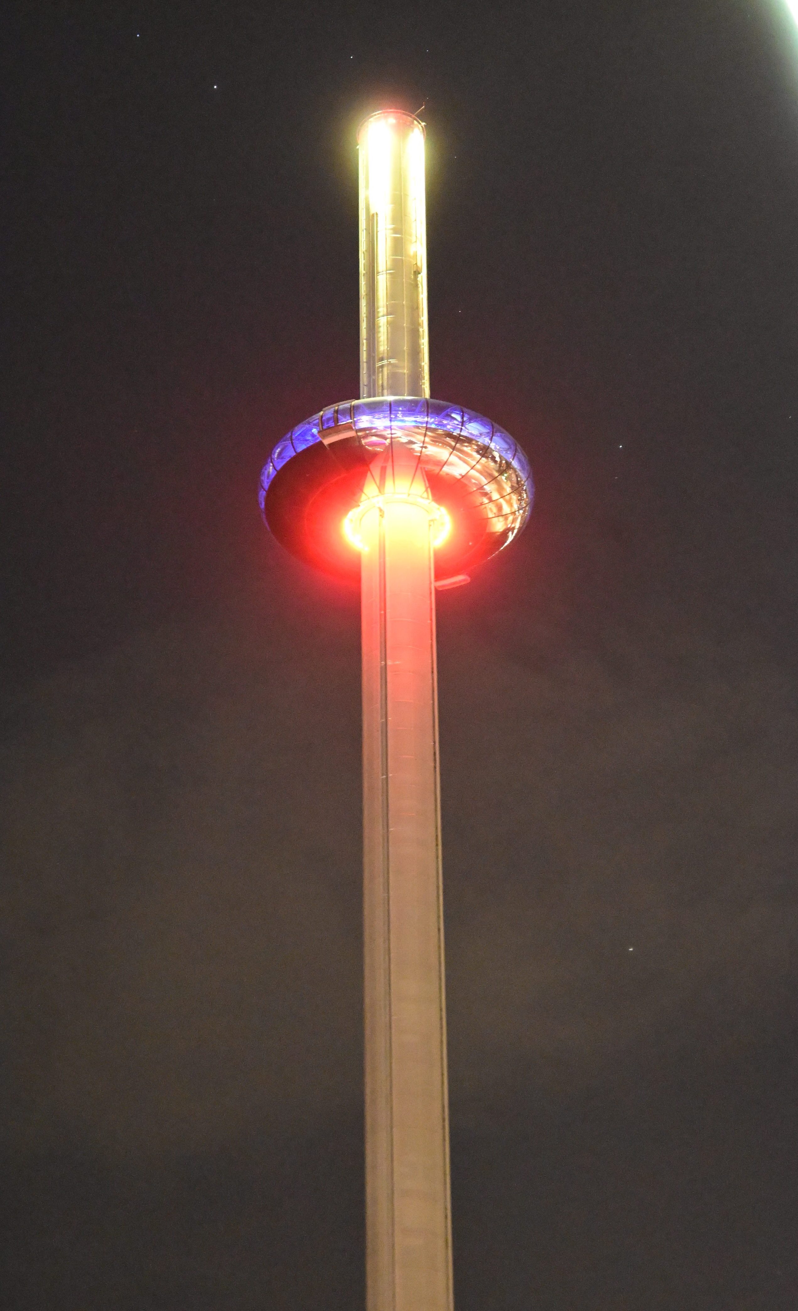 A tall observation tower illuminated at night, featuring a glowing sphere at the top surrounded by coloured lights.